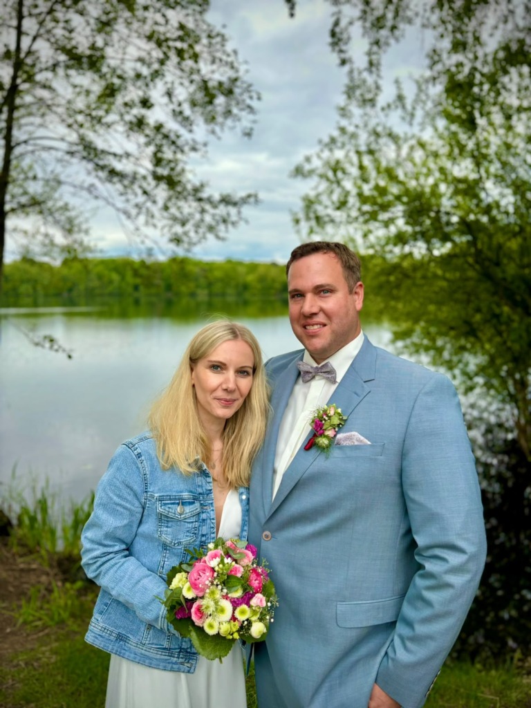 Indra und Daniel stehen mit Brautstrauss am Ufer eines Sees
